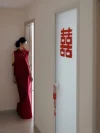 2 Piece Red Chinese Cheongsam - Bride in modern two-piece cheongsam dress, leaning against a wall in a wedding hallway with red double happiness sign on the door, showcasing the form-fitting silhouette of the red sleeveless top and maxi skirt.