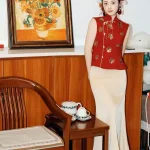 2 Piece Red and Gold Qipao - Chinese modern qipao with gold floral sequins on red sleeveless vest, paired with cream satin maxi skirt, posing in cozy home setting with teaware and sunflower painting