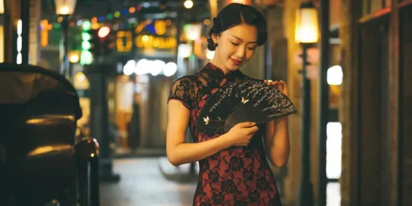 Woman wearing elegant red and black lace cheongsam dress, holding a decorated folding fan in a warm neon-lit vintage night Shanghai street, embodying classic 1930s Chinese qipao glamour.