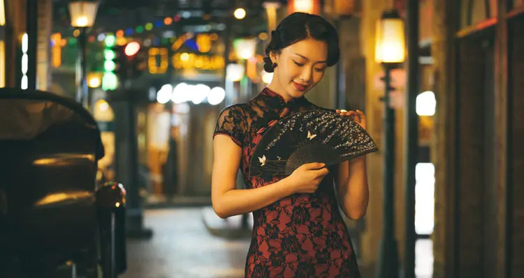 Woman wearing elegant red and black lace cheongsam dress, holding a decorated folding fan in a warm neon-lit vintage night Shanghai street, embodying classic 1930s Chinese qipao glamour.