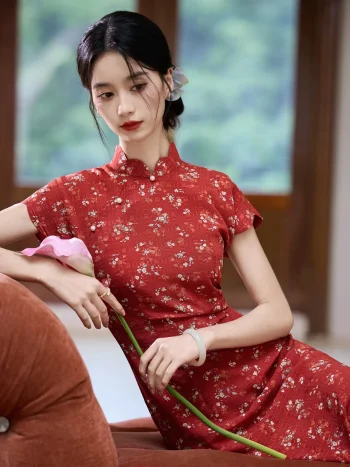 Close-up shot of a model in a red floral Chinese qipao dress, leaning on a sofa and holding a pink lily, highlighting the fabric texture and the dress's mandarin collar and buttons.
