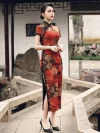 Profile view of a woman in a red floral vintage silk qipao dress standing on a stone bridge, highlighting the high side slit, lace trim, and traditional frog buttons along the placket.