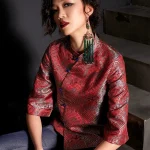 Close-up shot of a woman wearing the modern Chinese red jacquard satin cheongsam top, featuring intricate wave pattern, blue frog buttons, and high collar; paired with jeans for contemporary qipao top fashion