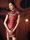 Model posing with hands clasped in a red modern mini qipao dress, highlighting the classic mandarin collar, green frog buttons, chest cutout, and elegant wave brocade pattern in a warm-toned studio setting.