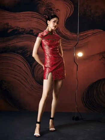 Full-body studio shot of model in a form-fitting red modern design short qipao dress, featuring a high front slit, sleek silhouette, and styled with strappy heels against a textured warm background.