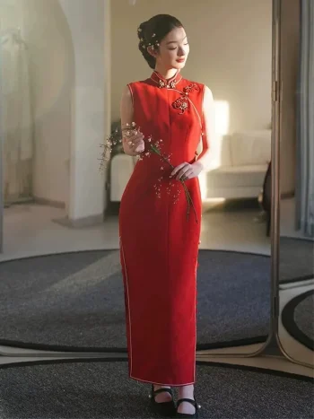 Red Modern Modified Sleeveless Long Wedding Qipao Dress - Bride in elegant sleeveless qipao wedding dress, standing in a sunlit trial room in front of a mirror, holding a delicate flower branch. The dress features intricate floral embroidery at the mandarin collar, form-fitting silhouette, and side slits, perfect for modern Chinese wedding ceremonies and photoshoots.