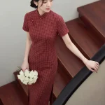 Women's 1940s traditional red plaid qipao, a classic red qipao dress with a refined checkered pattern, tailored fit, and vintage-inspired design. The model stands on a wooden staircase holding a bouquet of white roses, showcasing the flattering silhouette and elegant charm of this timeless traditional qipao.