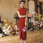 Red Silk Cheongsam for Wedding - Bride in elegant modern red silk cheongsam holding pink calla lilies at a Chinese wedding ceremony, wearing form-fitting sleeveless qipao with mandarin collar, surrounded by floral decorations.