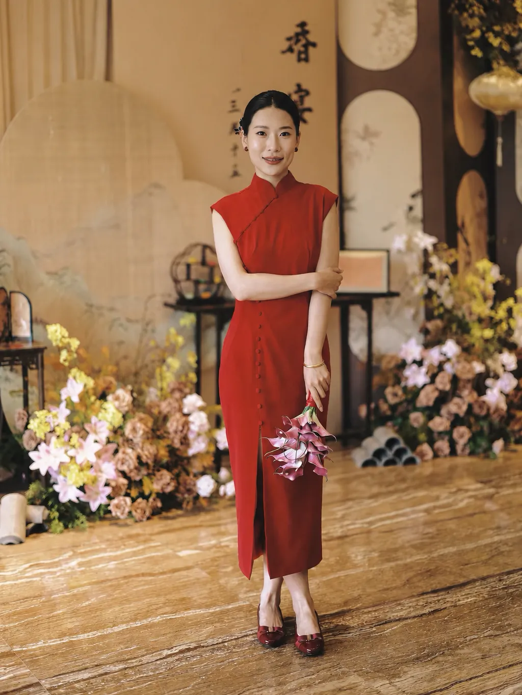 red-silk-cheongsam-for-wedding-calla-lilies-wedding-ceremony Red Silk Cheongsam for Wedding - Bride in elegant modern red silk cheongsam holding pink calla lilies at a Chinese wedding ceremony, wearing form-fitting sleeveless qipao with mandarin collar, surrounded by floral decorations.