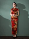 Full-body shot of a woman in a red floral pattern long silk qipao, holding a teacup against a muted green wall, showcasing the form-fitting cut, high slit, and luxurious mulberry silk sheen.