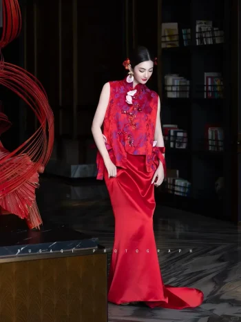 Red Silk Qipao Set - Woman in modern red qipao set with brocade vest and trailing satin mermaid skirt, standing in a luxury hotel lobby with a bookshelf backdrop, elegant two-piece chinese qipao for wedding photoshoots