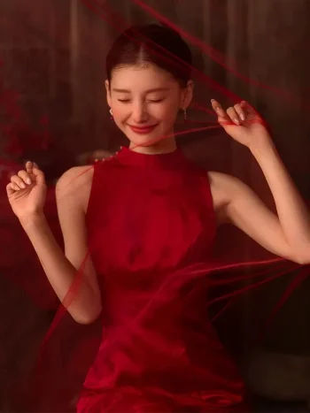 A bride in a red wedding cheongsam playfully interacts with red tulle, showcasing the sleeveless high-neck front of this modern Chinese style wedding dress. The hidden backless design adds a romantic, contemporary twist to the classic cheongsam silhouette.