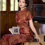 Full-body view of a woman sitting on a wooden chair, wearing a 1930s reddish-brown floral silk qipao and holding a book, showcasing the elegant silhouette and vintage charm of the traditional Chinese dress in a warm indoor setting.