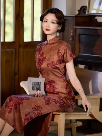 Full-body view of a woman sitting on a wooden chair, wearing a 1930s reddish-brown floral silk qipao and holding a book, showcasing the elegant silhouette and vintage charm of the traditional Chinese dress in a warm indoor setting.