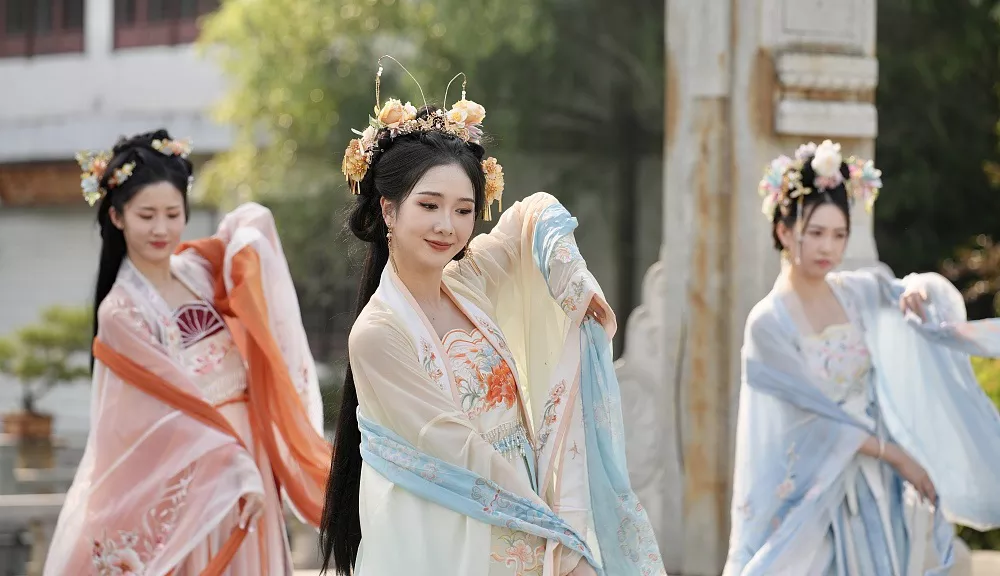 Three women wearing elegant Tang-style traditional Chinese hanfu dresses with sheer embroidered sleeves and floral hair ornaments, performing graceful movements in a classic Chinese garden setting.