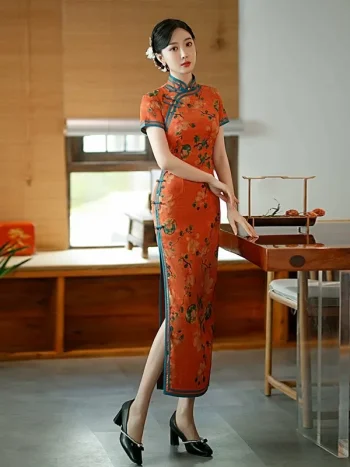 Old Shanghai inspired long cheongsam in tangerine red with peony floral print and blue trim, women's traditional Chinese dress with high slit, model leaning on table.