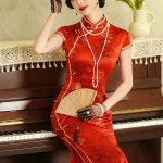 Model wearing a traditional Chinese wedding red long qipao, seated on a vintage piano with a folding fan, showcasing the elegant satin fabric and delicate floral print.