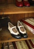 Two-tone black and white Margarita 1920s vintage schoolgirl leather Qipao shoes with rivet accents, displayed on a wooden shelf alongside other vintage footwear styles.