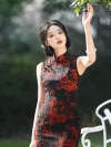 Close-up of women's vintage floral sleeveless high cut cheongsam with red dragon print on black fabric, model raising hand to touch tree leaves outdoors, showing fabric texture and print details.