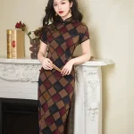 Women's vintage plaid short sleeve cheongsam with a high slit, featuring a bold geometric checkered pattern in brown, red, and blue. The model leans on a white fireplace mantel holding a folding fan, highlighting the dress’s elegant silhouette and timeless design.