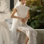 Vintage Modified White Lace Wedding Qipao Dress - Bride sitting in a traditional Chinese garden, wearing a short-sleeve white lace qipao with floral headpiece and lace veil, holding a hand fan, showcasing the lace texture and high collar with pearl details, ideal for elegant bridal portraits.