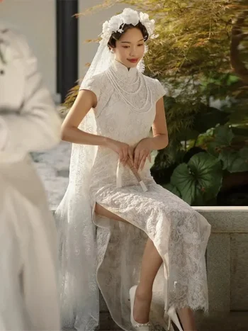 Vintage Modified White Lace Wedding Qipao Dress - Bride sitting in a traditional Chinese garden, wearing a short-sleeve white lace qipao with floral headpiece and lace veil, holding a hand fan, showcasing the lace texture and high collar with pearl details, ideal for elegant bridal portraits.
