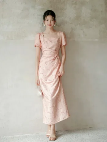 Wall-leaning view of women's summer modified cheongsam, new Chinese style long dress with square neck puff sleeve, waist cinched pleated design in soft pink textured fabric showing elegant drape