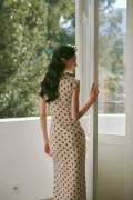 Back view of a woman in the "Waltz of Youth" midcentury Hong Kong style cotton qipao dress, standing by a sunlit window. The image highlights the elegant form-fitting silhouette, classic polka dot print, and cap sleeve design of this daily wear cheongsam.