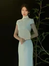 White Lace Fishtail Wedding Cheongsam Dress - Elegant woman in a white lace cheongsam wedding dress, standing against a dark background with bamboo decor, holding a lace fan. The dress features a high mandarin collar, short cap sleeves, intricate lace texture, and a form-fitting fishtail silhouette, perfect for bridal photoshoots and modern Chinese weddings.