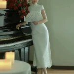 White Lace Fishtail Wedding Cheongsam Dress - Full-body shot of a woman in a white lace fishtail cheongsam wedding dress, leaning against a grand piano with red roses and candles. The lace cheongsam features a form-fitting silhouette, lace trim, short sleeves, and side slit, creating a romantic bridal look for wedding day and formal events.