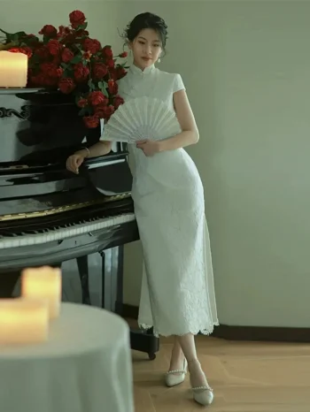 White Lace Fishtail Wedding Cheongsam Dress - Full-body shot of a woman in a white lace fishtail cheongsam wedding dress, leaning against a grand piano with red roses and candles. The lace cheongsam features a form-fitting silhouette, lace trim, short sleeves, and side slit, creating a romantic bridal look for wedding day and formal events.