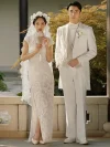 White Lace Qipao Wedding Dress - Full-body shot of bride in a vintage modified white lace qipao, standing beside her groom in a white suit in a traditional Chinese setting, holding a hand fan, featuring a side slit and intricate lace texture for a classic bridal look.
