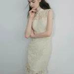 Elegant woman in a white modern stylish Chinese short cheongsam dress, posing with a thoughtful look against a plain background, showcasing the intricate lace texture, form-fitting silhouette, and delicate ruffled hem.