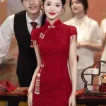 Bride wearing a wine red floral lace chinese wedding cheongsam dress, elegant midi cheongsam with traditional tassel and pearl details, posing at a classic chinese wedding ceremony.