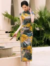 Model holding a hand fan, wearing a women high end yellow mulberry silk cheongsam dress with landscape print, posing near tropical plants for a vintage elegant look.