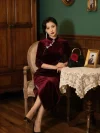 Gorgeous asian lady dressed in premium Long Sleeve Cheongsam crafted from burgundy velvet fabric, resting arms on lace covered table with rose decorated handbag and vintage photo frames beside antique wooden cabinet, timeless elegant Cheongsam Velvet banquet dress