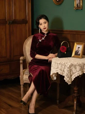 Gorgeous asian lady dressed in premium Long Sleeve Cheongsam crafted from burgundy velvet fabric, resting arms on lace covered table with rose decorated handbag and vintage photo frames beside antique wooden cabinet, timeless elegant Cheongsam Velvet banquet dress