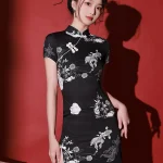 Woman modeling a black mini cheongsam dress with white floral and fish embroidery in a red studio.