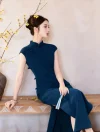 The seated view of a casual blue qipao outfit, model relaxing on a wooden bench, holding a folding fan beside blossoming yellow branches.