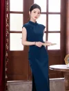 A graceful woman wearing a sleeveless blue qipao dress with traditional knot buttons, holding a book near a vintage window and curtains.