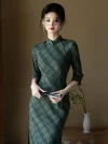 Elegant woman wearing a cheongsam dress long sleeve in olive green plaid, holding a black folding fan, standing gracefully in a minimalist room with a white vase.