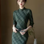 Elegant woman wearing a cheongsam dress long sleeve in olive green plaid, holding a black folding fan, standing gracefully in a minimalist room with a white vase.