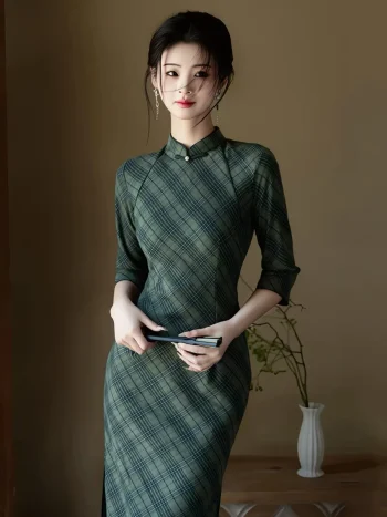 Elegant woman wearing a cheongsam dress long sleeve in olive green plaid, holding a black folding fan, standing gracefully in a minimalist room with a white vase.