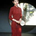 Elegant bride in a red long sleeve bridal qipao with intricate floral jacquard, holding a round white fan in front of a traditional Chinese moon gate.