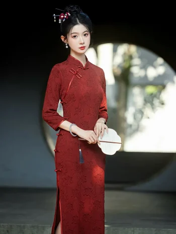 Elegant bride in a red long sleeve bridal qipao with intricate floral jacquard, holding a round white fan in front of a traditional Chinese moon gate.