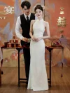Stunning full-body shot of a modern white cheongsam featuring a sleek halter neckline, worn by a bride participating in a traditional red tea ceremony with a groom standing behind.