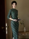 Beautiful woman in a fitted long sleeve cheongsam dress featuring olive green plaid and side slits, posing gracefully near a wooden table displaying a minimalist vase arrangement.