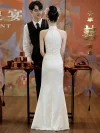 Back view of an elegant woman in a long white halter cheongsam showing the elegant cut of the back and the flowing dress silhouette, standing in a vintage Oriental-style event hall.