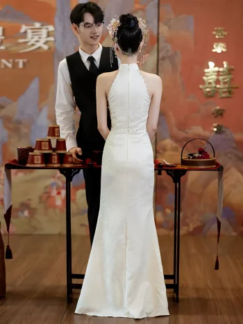 Back view of an elegant woman in a long white halter cheongsam showing the elegant cut of the back and the flowing dress silhouette, standing in a vintage Oriental-style event hall.
