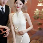 Elegant bride wearing a white halter cheongsam with delicate pearl decorations and a traditional floral headpiece, standing gracefully next to a groom in a black vest during a Chinese wedding.